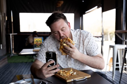Nick takes a bite out of a massive sandwich while filming himself on his phone