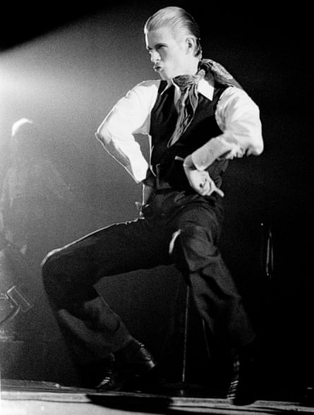 David Bowie poses dramatically on stage in a waistcoat with a scarf tied at his neck
