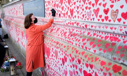 The National Covid Memorial Wall opposite Westminster: a campaigner, wearing a black face mask, writes in one of the red hearts that have been painted on a long stretch of pale stone wall. She is wearing a red coat, and flowers and a lantern are seen behind her.