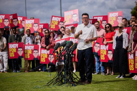 Burnham speaks with microphones in front of him and dozens of supporters behind, with placards of the illustration by Stanley Chow
