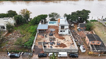 Seen from above, a large house with a pool being built by a river. Around it is rubble and devastation.