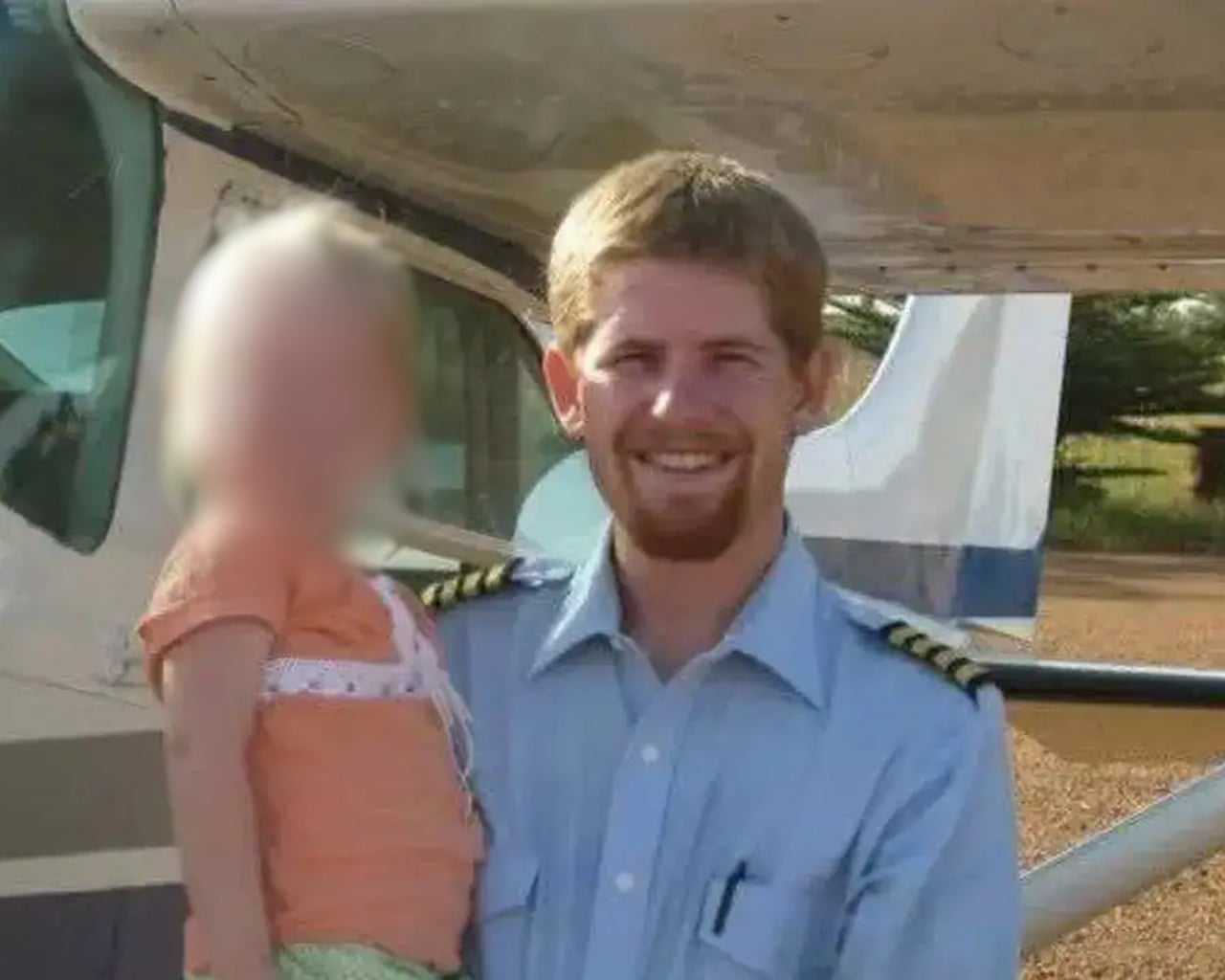 Kevin Rideout wearing a pilot’s uniform, holding a child with blurred-out face while standing outside a plane