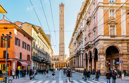 A wide, sunlit street in Bologna with Asinelli tower in the centre, and people using a pedestrian crossing.