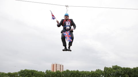 Boris Johnson hanging on a zip wire holding union jack flags over a park, with a tower block in the background