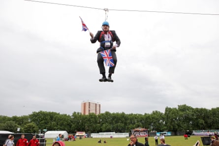 Boris Johnson hanging on a zip wire holding union jack flags over a park, with a tower block in the background 