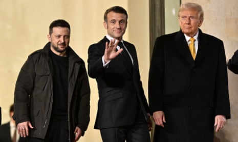 Zelenskyy, Macron and Trump stand together outside the French government building. Macron is waving to the camera in the centre; he and Trump wear shirts and ties but Zelenskyy wears an anorak and black jumper