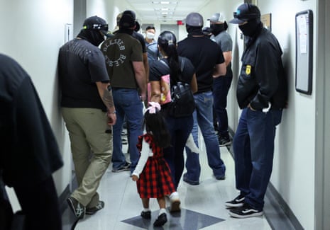 A small girl in a plaid dress walks past masked federal agents lining a courthouse hallway