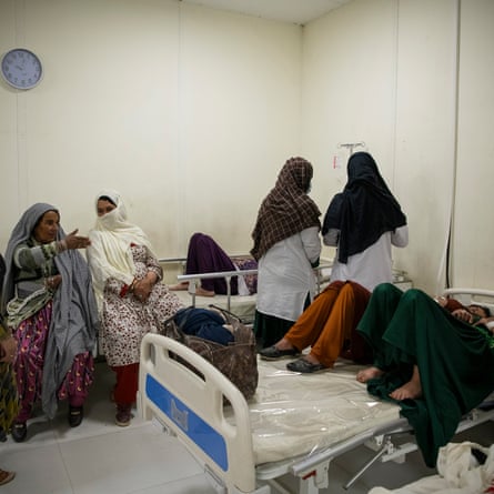 A busy maternity ward where two women in the foreground share one bed, while two female nurses stand next to a patient in a second bed, with two older women sitting on the end of it, chatting