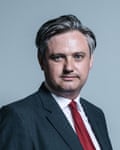 John Woodcock: official head and shoulders portrait. He is in his mid-to-late 40s with grey hair swept back from his forehead and has a round face; he wears a dark grey jacket, white shirt and red tie.