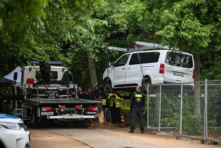 A white Citroën passenger van is lifted over wire fencing towards a tow truck on a tree-lined path as police stand by.