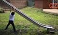 A pupil hangs off the side of a slide on patchy grass with a safety barrier by a wooden shed in the background