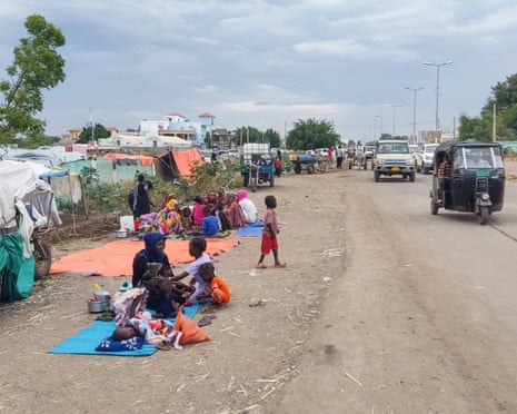 Displaced families sit on blankets beside a road near Gedaref, Sudan, with tents and vehicles nearby