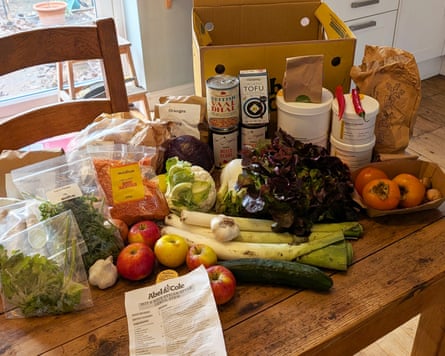 The contents of an Abel and Cole recipe box is displayed on a wooden dining table. There is a selection of fresh fruit and veg, tinned goods and herbs in bags.