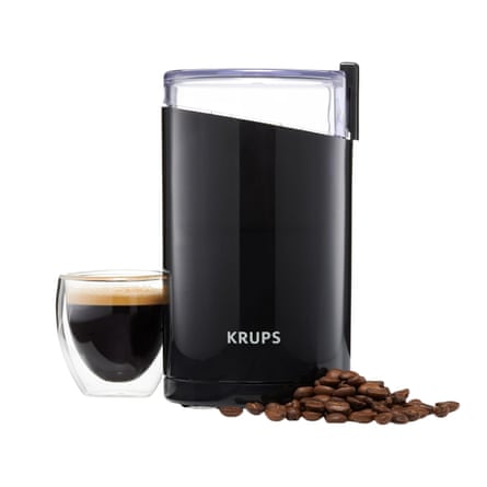 A black Krups coffee grinder stands beside an espresso cup and scattered coffee beans