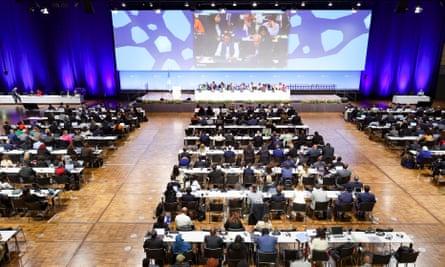 General view of people seated in a conference hall facing a panel of experts, as a big screen shows proceedings behind