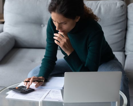 A woman sits on a sofa using a calculator while looking at papers, with a laptop nearby