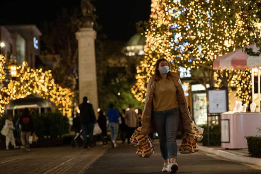 A woman wearing a mask carries shopping bags through an outdoor shopping center at night. The trees in the background are covered in tiny lights.