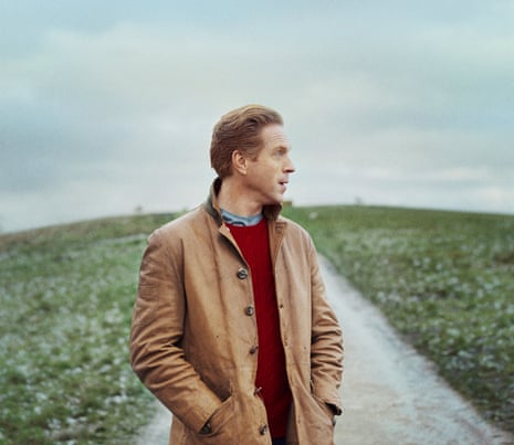 A portrait of Damian Lewis standing on a road on a hill, wearing a red jumper and tan coat