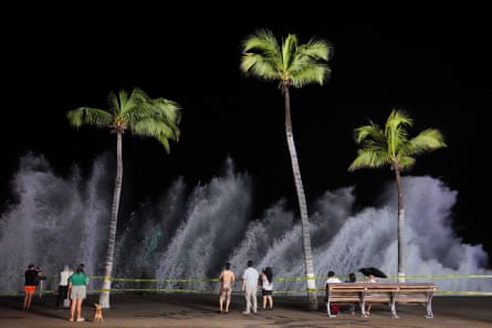 People stand on a promenade between tall palm trees as high waves crash in front of them.