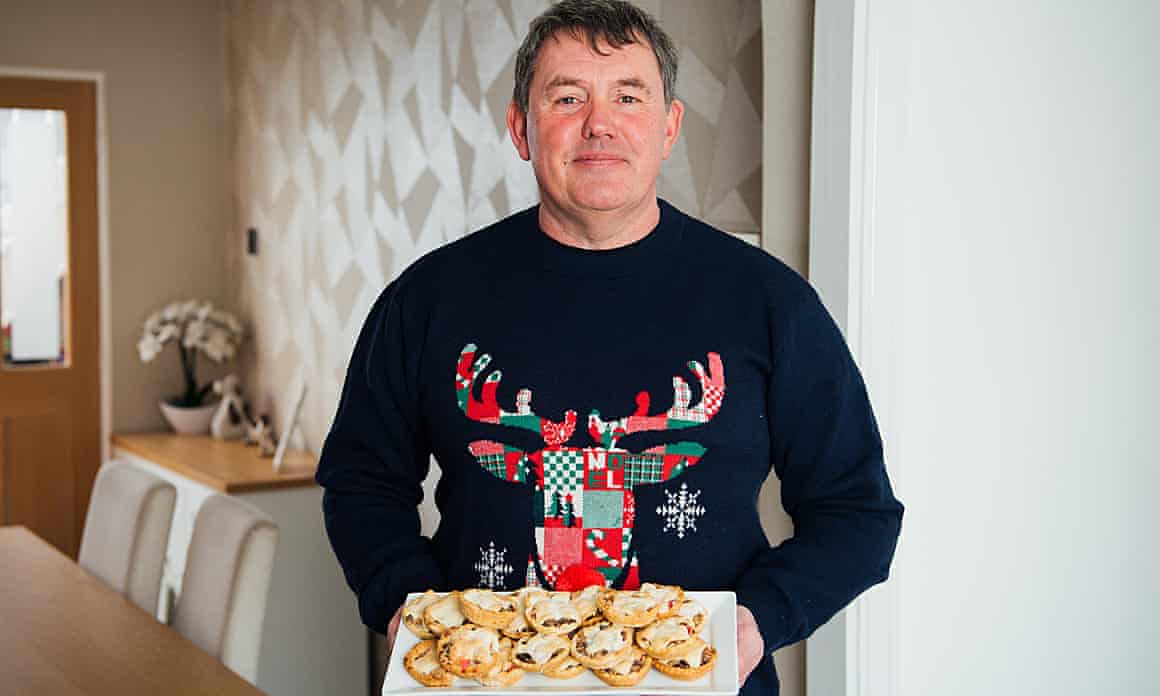 A man in a Christmas jumper depicting a patchwork reindeer holds a tray of mince pies