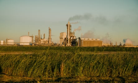 In an early dawn, seen from across a tall gree field, is a plant with smokestacks chugging smoke into the sky.