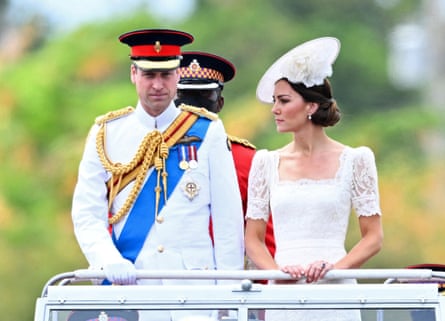 William in full uniform and the Duchess in a white dress and hat standing in the back of an open-topped vehicle