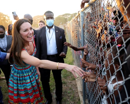 The Duchess of Cambridge smiles while reaching through a chain-link fence to touch children’s hands.