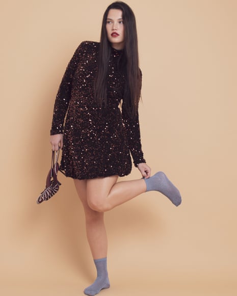 Model with long grown hair wearing short brown glittery dress and purple socks, and holding sparkly shoes
