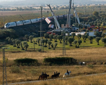 Cranes working at the derailed train crash site as people on horses ride past