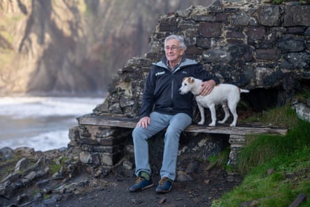 Peters sat smiling with a dog on a bench, rocks and the sea in background