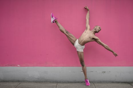 Dancer Juan Pablo Rodríguez stands in a very balletic pose wearing only shorts and trainers while in front of a mainly pink background