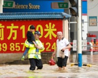 A police officer in a reflective vest carries a child through knee-deep flood water on a city street