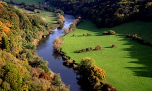 A meandering river bordered by woods in autumn colours and green fields