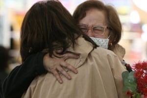 A woman is wearing a mask and glasses and holding flowers while she hugs another woman who has long dark hair and is wearing a beige coat. Sisters Lina and Adeline embrace as they reunite at Sydney’s international airport