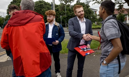 Two young men with red hair and beards, the candidate wearing a grey suit and white shirt, hold clipboards and Labour leaflets as they shake hands and talk to others, including a young man with a red Labour rosette pinned to his T-shirt