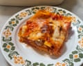 A square slice of lasagne on a patterned plate in front of a white oven dish containing the baked lasagne.