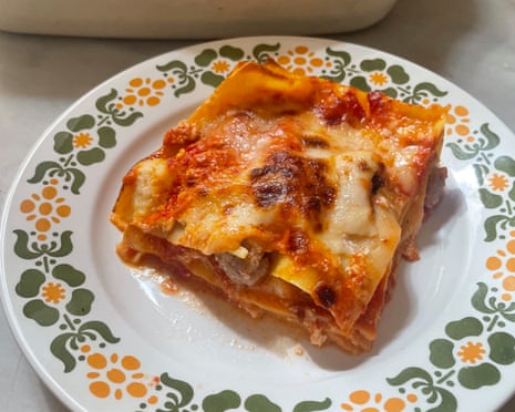 A square slice of lasagne on a patterned plate in front of a white oven dish containing the baked lasagne.