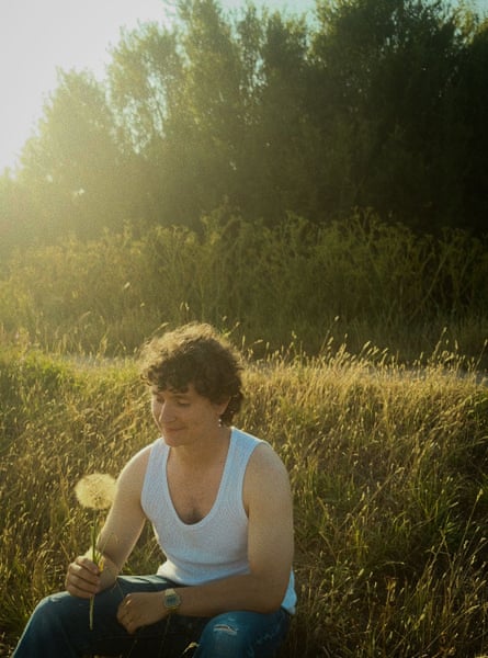 Browne sitting in a sunny field, wearing a white vest and a holding a dandelion seed head