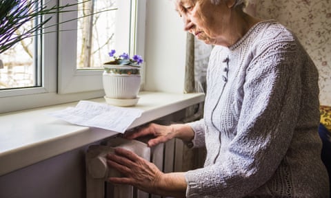 A senior woman holds a gas bill against the windowsill and puts her hands to a radiator; she is wearing a cardigan and the light suggests it is winter