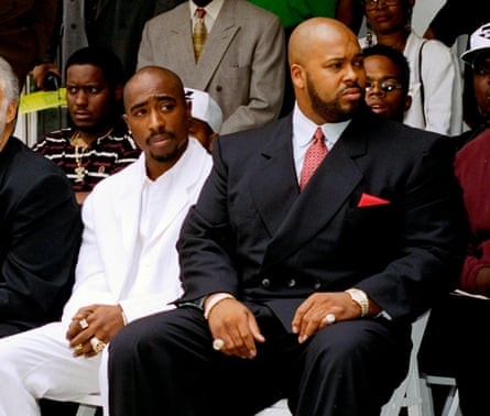 Seated on adjacent white folding chairs, amid rows of other people on white folding chairs, is a young Black man with a bald head and black mustache wearing a white suit, next to a much larger Black man, also with a bald head, who has a black beard and wears a black suit, light blue shirt, red tie, and red pocket square. The man’s hands rest on his thighs and showing two large pinky rings. 