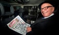 Murdoch seated in a car holding a newspaper and smiling to camera