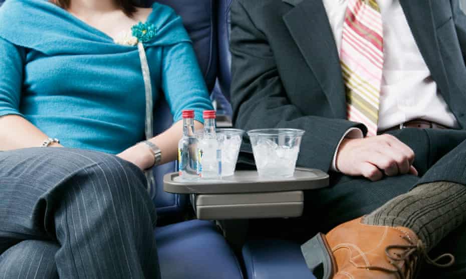 Two people, one wearing a blue top, the other a dark-grey suit and red-and-yellow stripy tie, sitting in airline seats with alcoholic drinks on a tray between them.