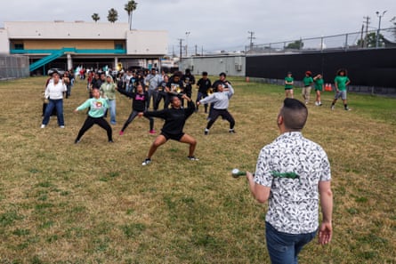 Man with baton under his arm faces teenagers dancing in formation on grassy lawn.