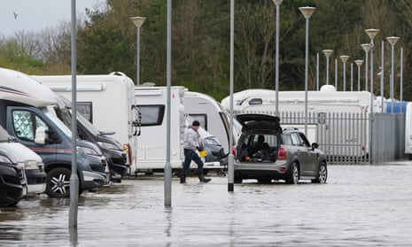 A man walks towards his car through ankle-high water. Caravans and motorhomes are parked behind