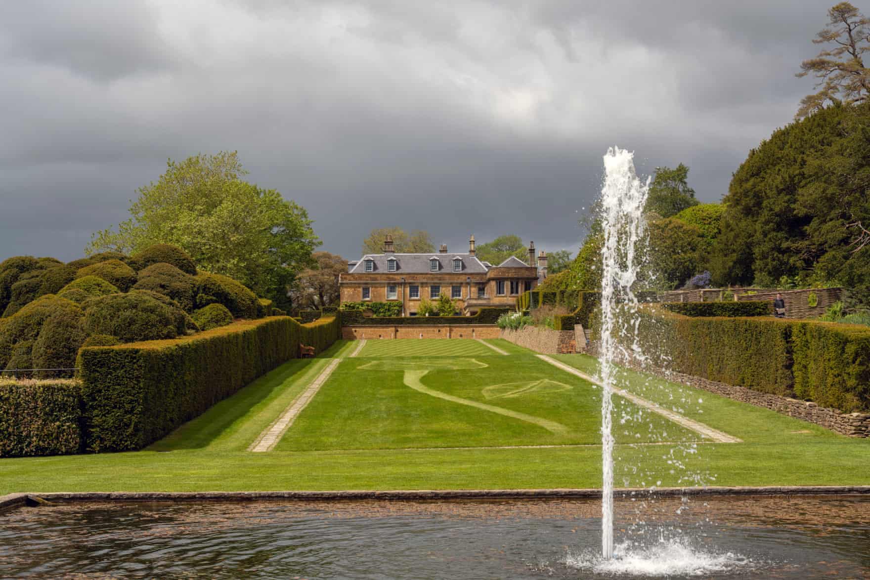 View of large country house across a manicured lawn with neat hedges to each side and a pool with spurting fountain in the foreground.