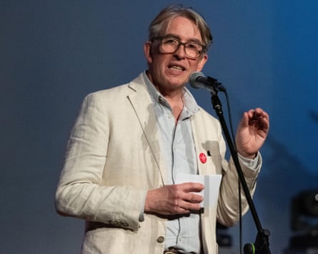 Steve Coogan, in glasses and a cream blazer, speaks into a microphone holding note cards