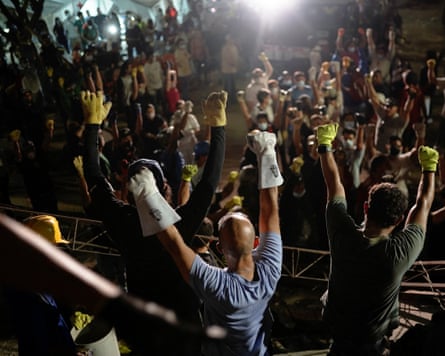 Rescue workers wearing gloves raise their fists above a large crowd of people, some of whom are doing the same