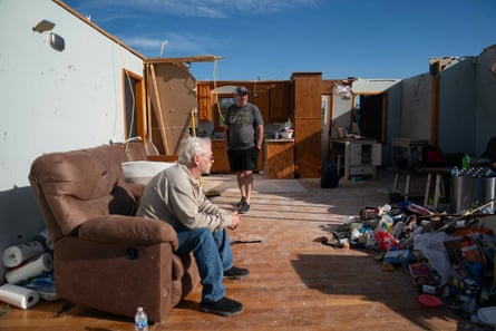 a man stands next to a man sitting in a house that had its roof turn off by a tornado