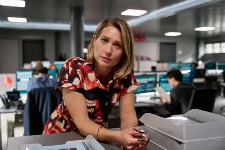 A woman in a red and cream patterned dress looks thoughtful as leans on a desk in an office