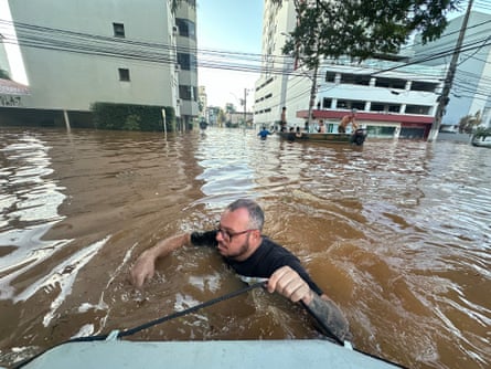 Up to his shoulders in flood water, a man pulls a boat in São Leopoldo, a suburb of Porto Alegre. There is another boat in the shot, with several people on board, navigating the flooded streets.